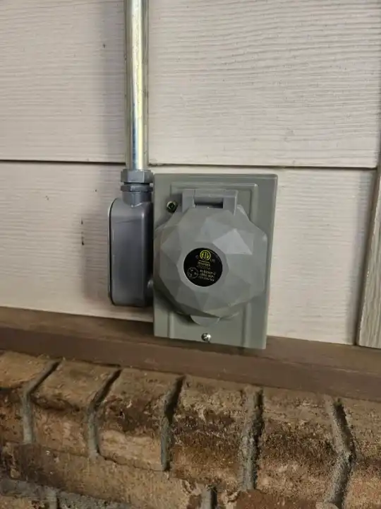 Gray outdoor receptacle mounted on beige house siding above a brick base, with metal conduit running up the wall beside it